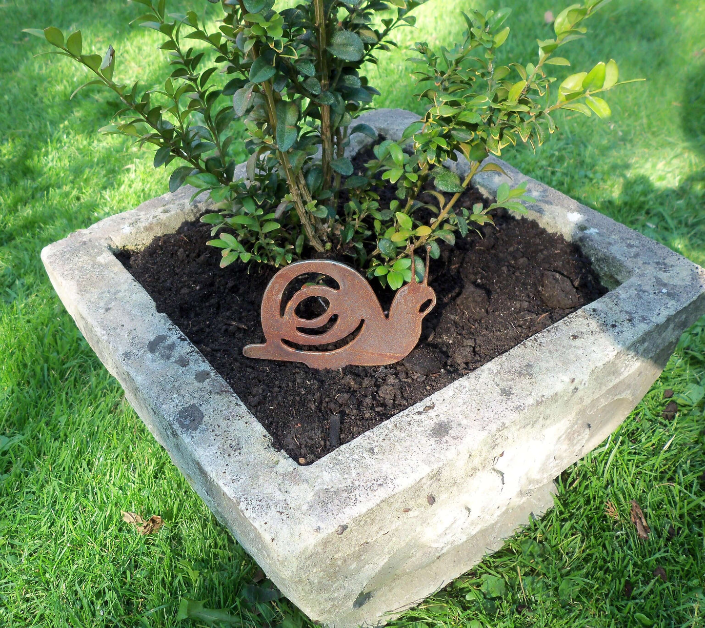 Rusty metal snail garden decoration with pot spike in a stone pot beside green plant, unique patina effect.