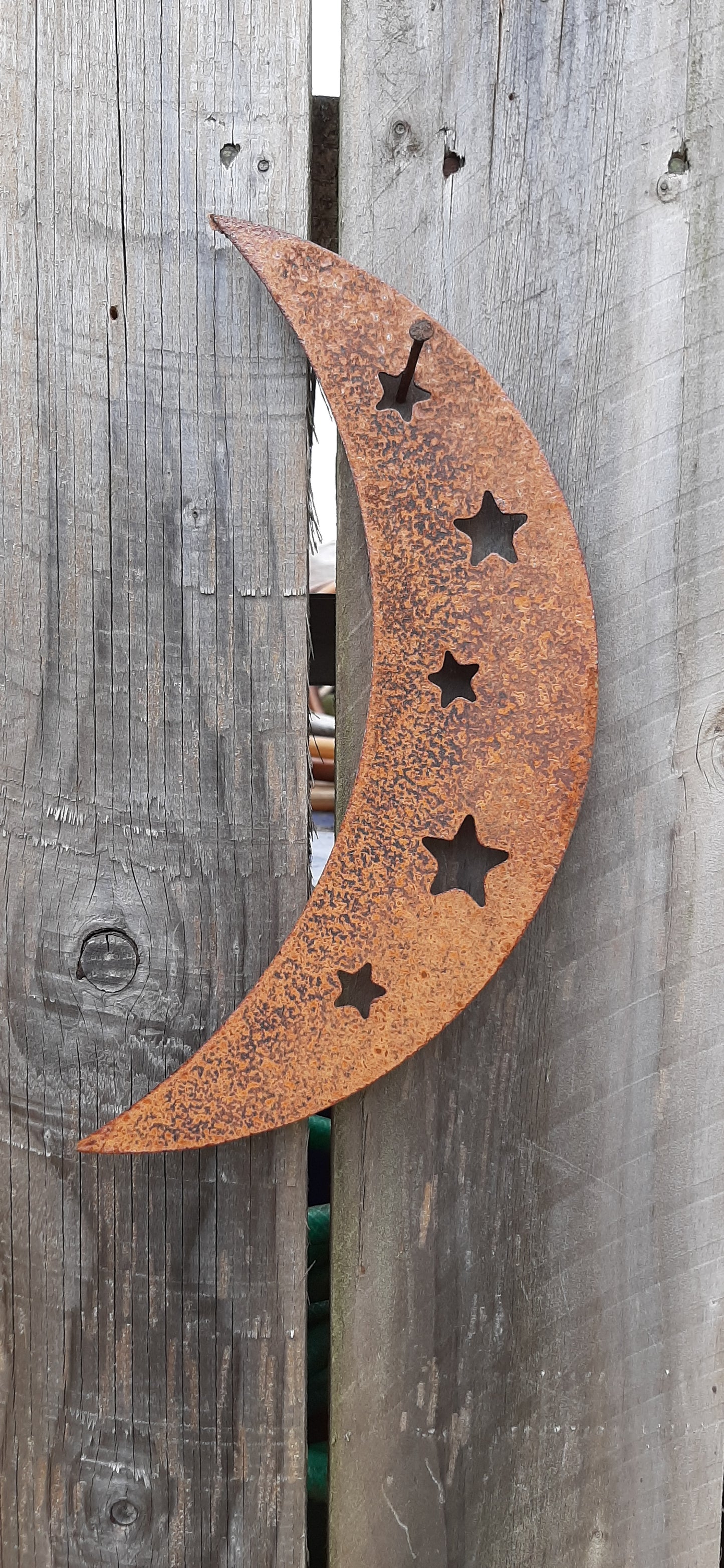 Large rustic metal crescent moon garden ornament with five star cutouts, hanging on wooden fence.
