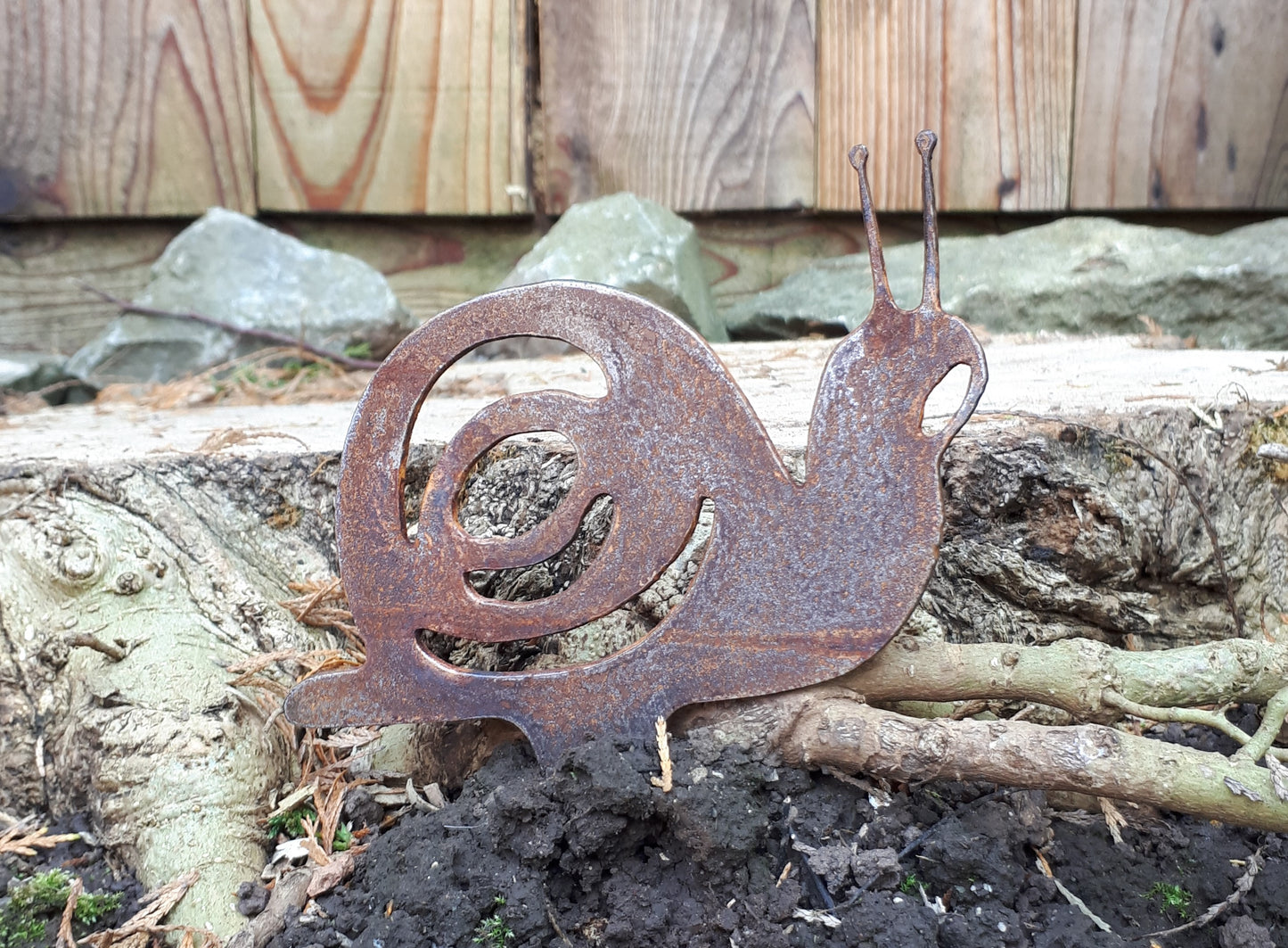 Rustic metal snail garden ornament with ground spike by a tree stump, unique patina effect.
