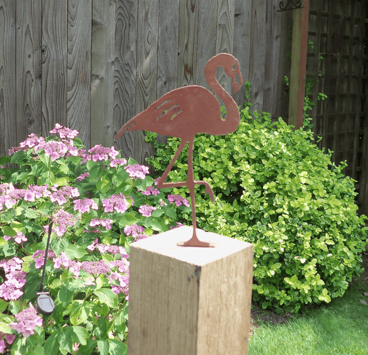 Rusty Metal Flamingo garden ornament displayed on a rustic wooden post surrounded by colorful garden flowers.