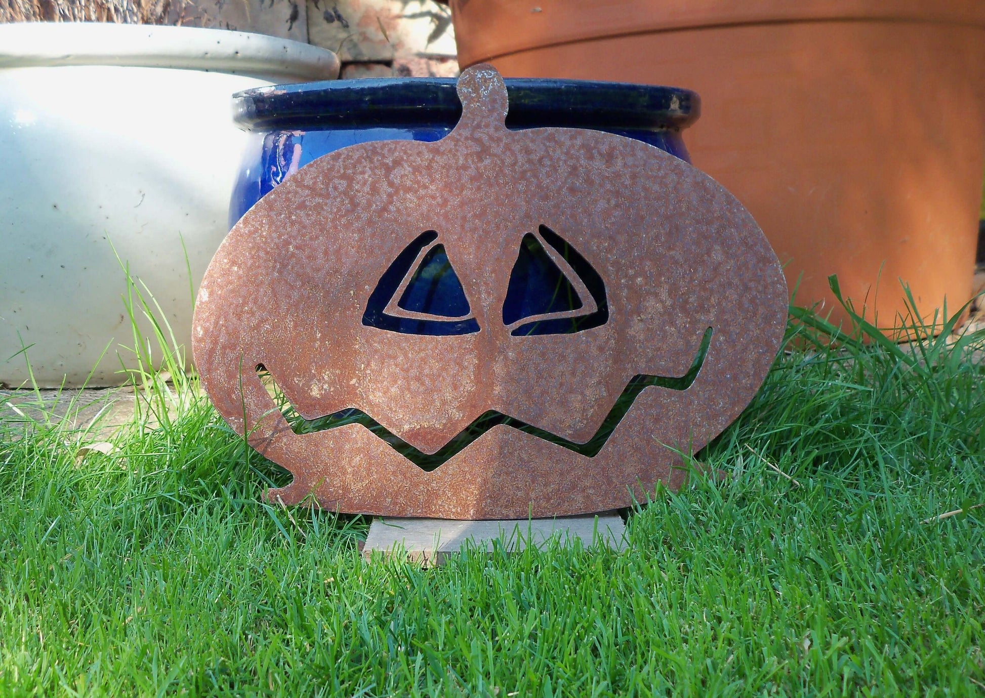 Rustic rusty metal pumpkin garden ornament against blue plant pot in green grass. Perfect Halloween garden decor.