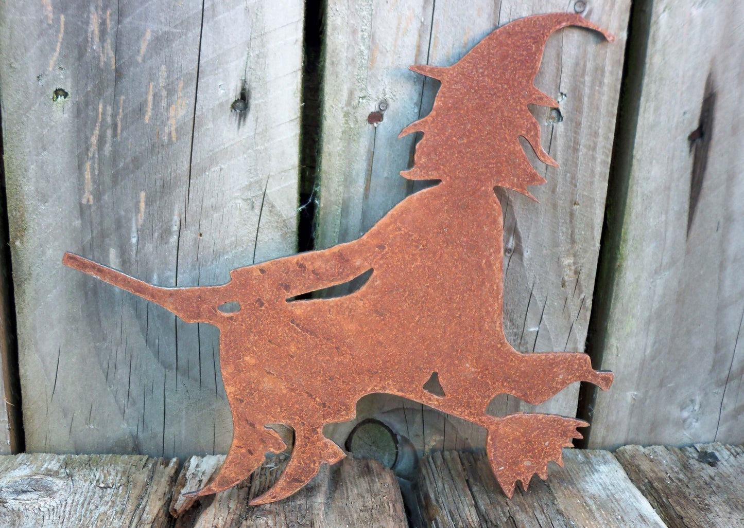 Rusty metal witch on broomstick garden ornament against wooden background, rustic Halloween decor.