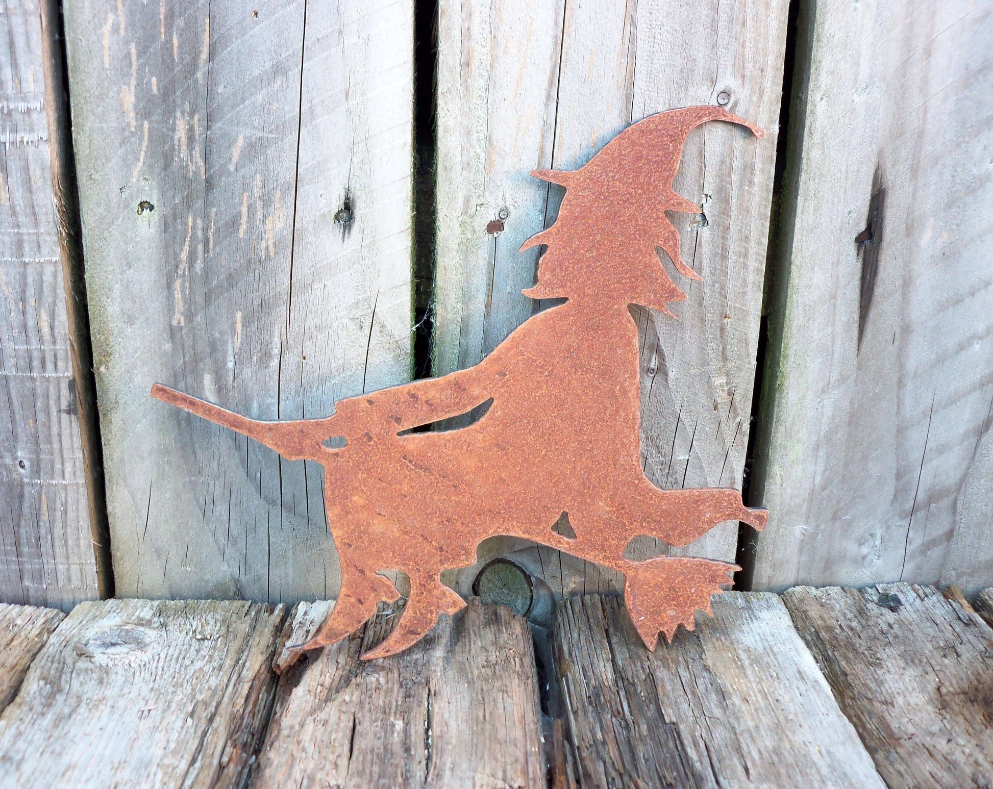 Rusty metal witch on a broomstick garden ornament against wooden background, rustic Halloween decor, 225mm x 265mm steel art.