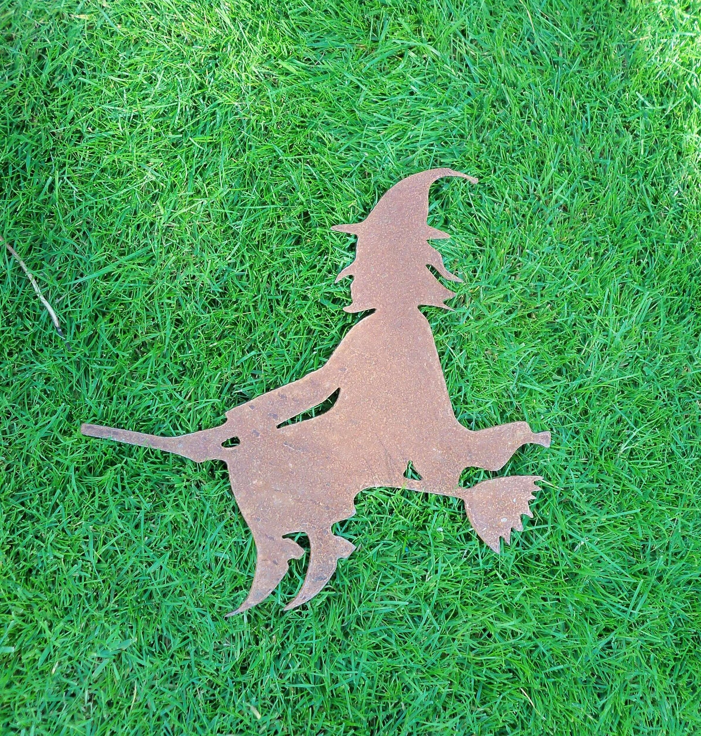 Rustic metal witch on broomstick garden ornament on grass, rusted steel design, perfect for Halloween and garden decor.