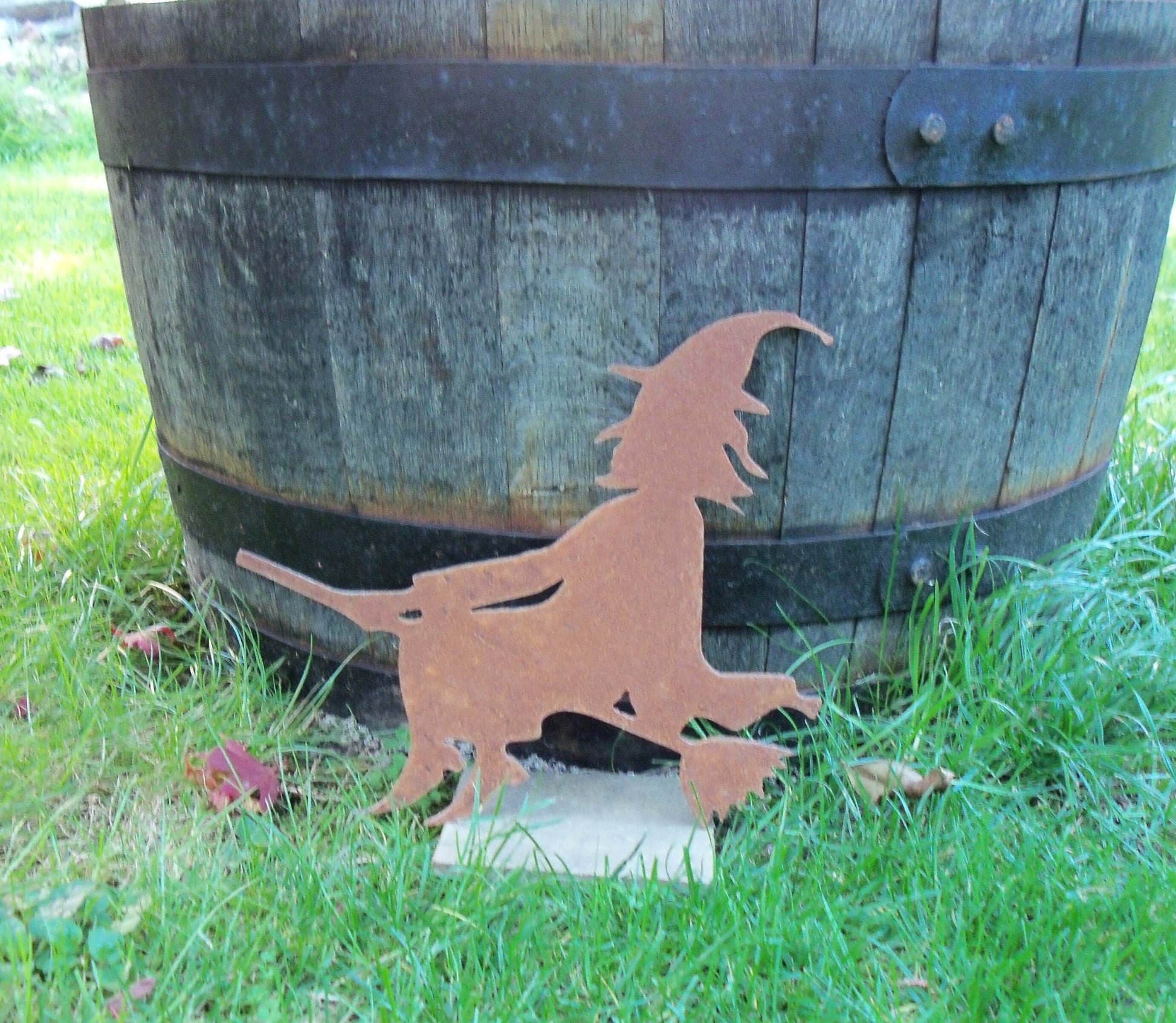 Rustic rusty metal witch on broomstick garden ornament against wooden barrel backdrop in green grass. Perfect Halloween garden decor.