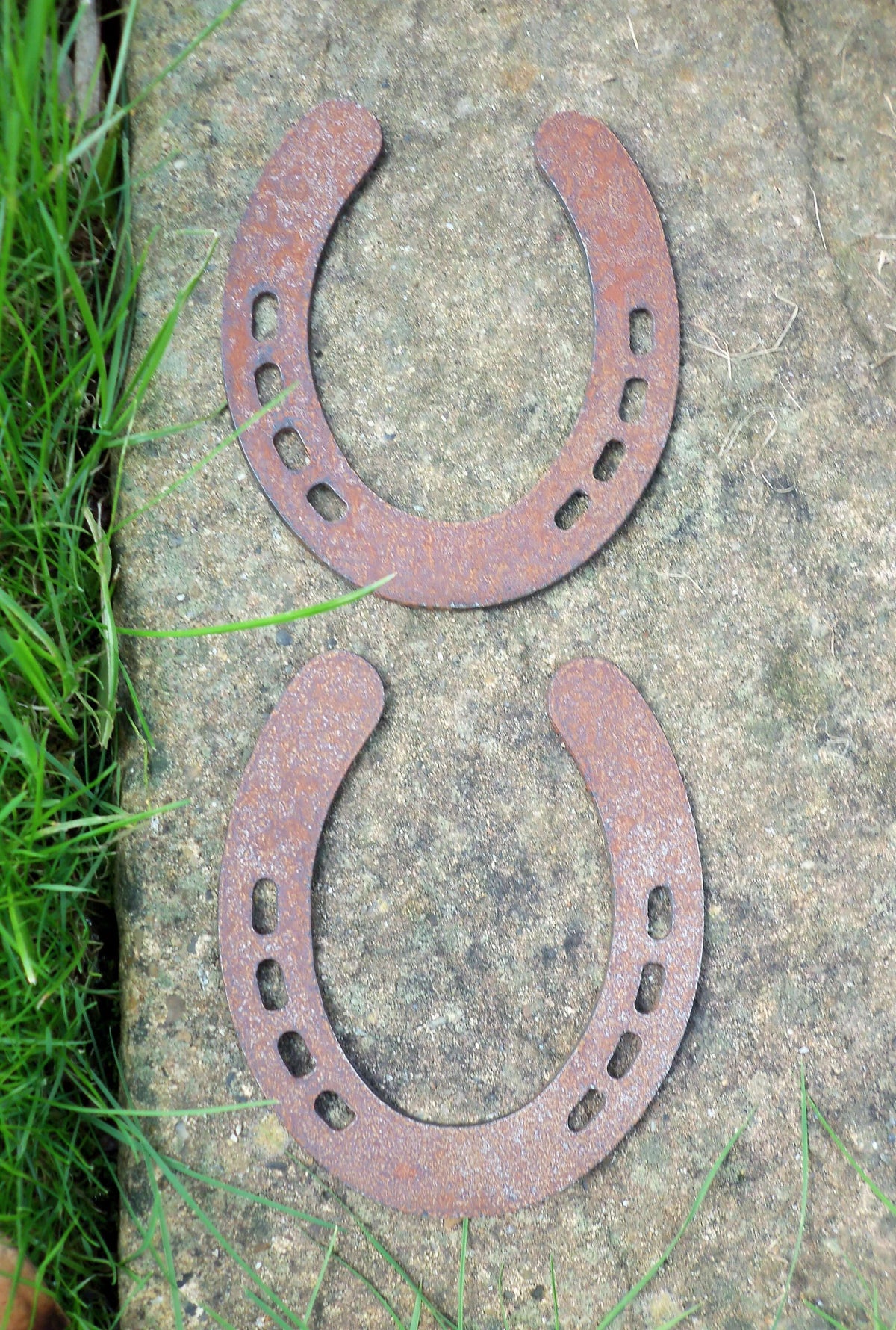 Rusty Steel PAIR OF HORSESHOES Garden Ornaments displayed on a stone surface surrounded by grass.