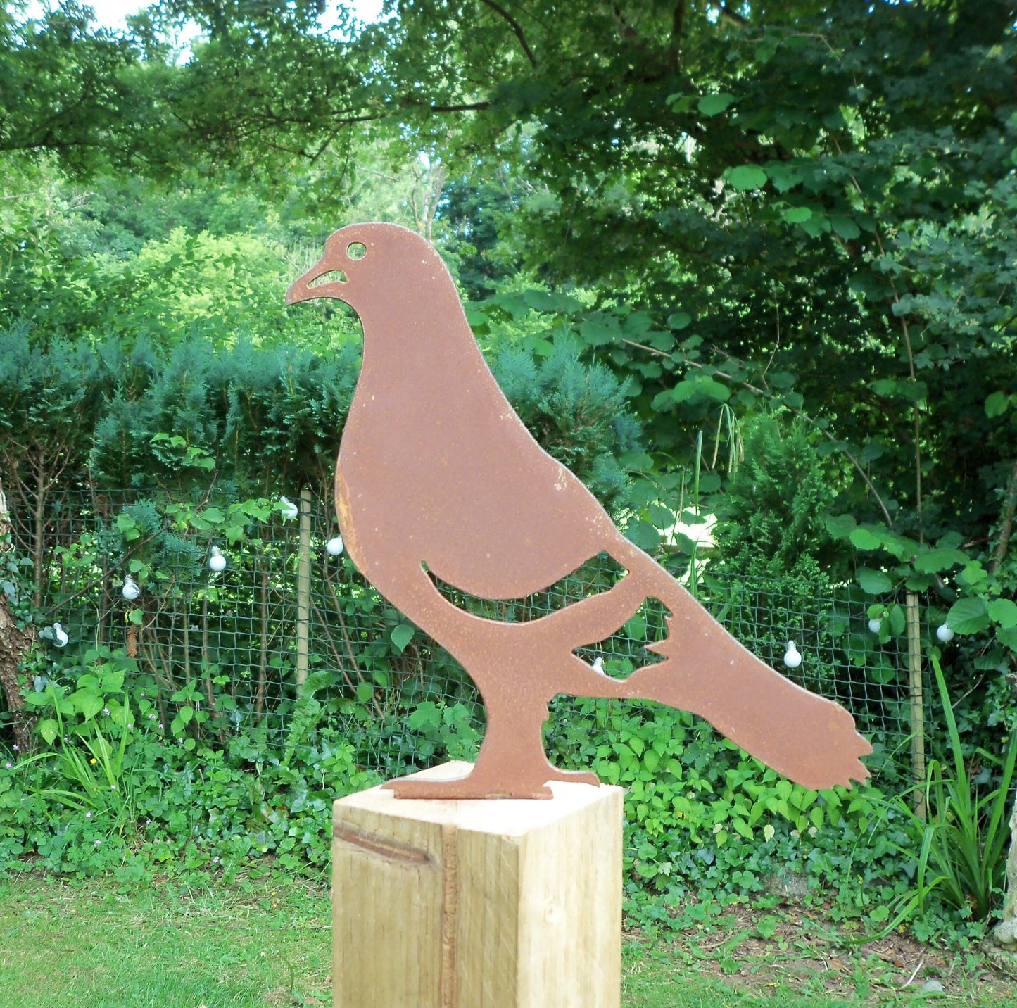 Rusty metal pigeon garden decor on a fence post besides trees and plants, Perfect garden feature.