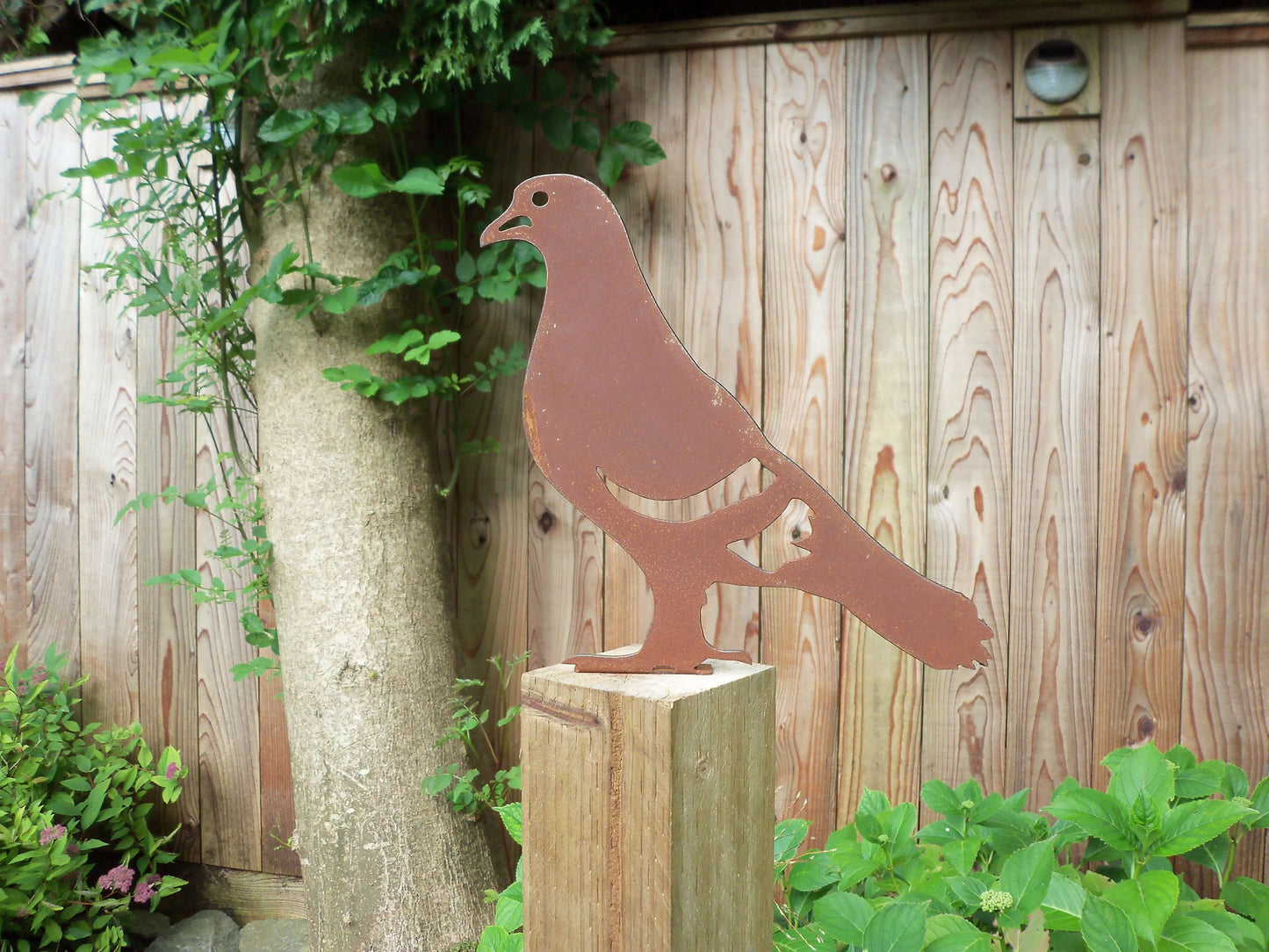 Rustic metal pigeon bird fence post topper garden decoration against a fence and tree trunk, unique rusted patina effect.