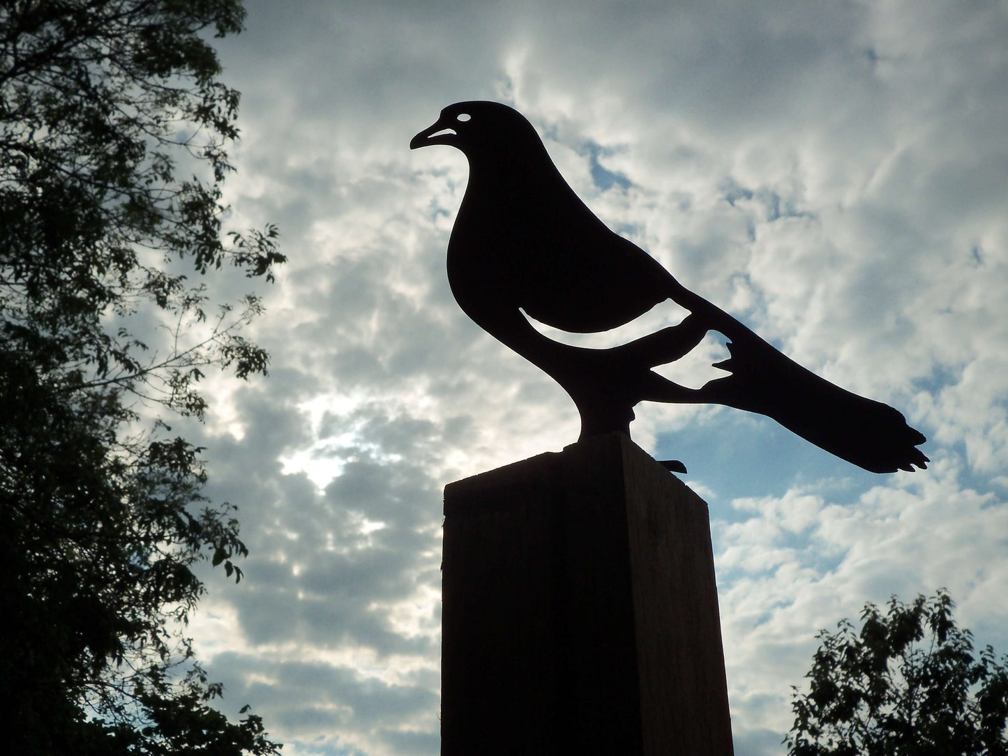 Rusty metal pigeon ornament on a post against a beautiful cloudy sky, ideal garden decoration.