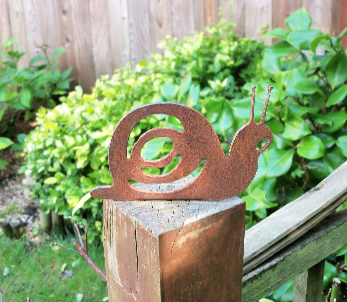 Rusty Steel Snail Garden Ornament on a wooden fence post in a garden setting, showcasing rustic metal decoration.