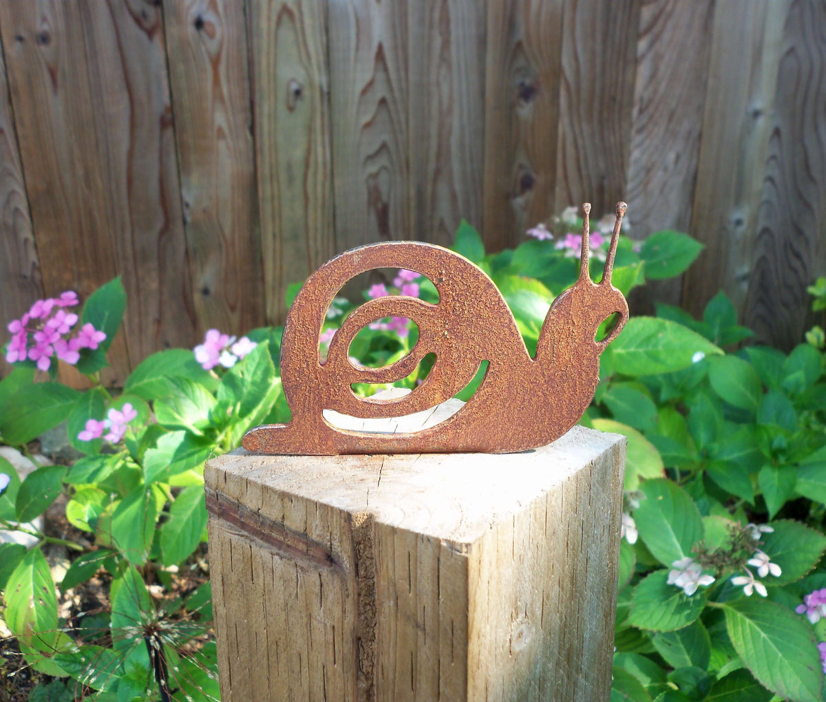 Rusty Steel Snail Garden Ornament placed on a wooden post surrounded by green plants and flowers.