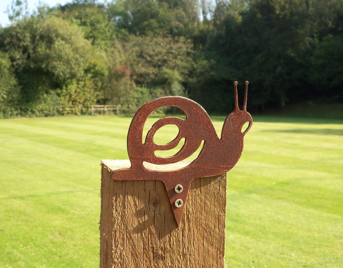 Rusty Steel Snail Garden Ornament on a fence post surrounded by a lush green garden.