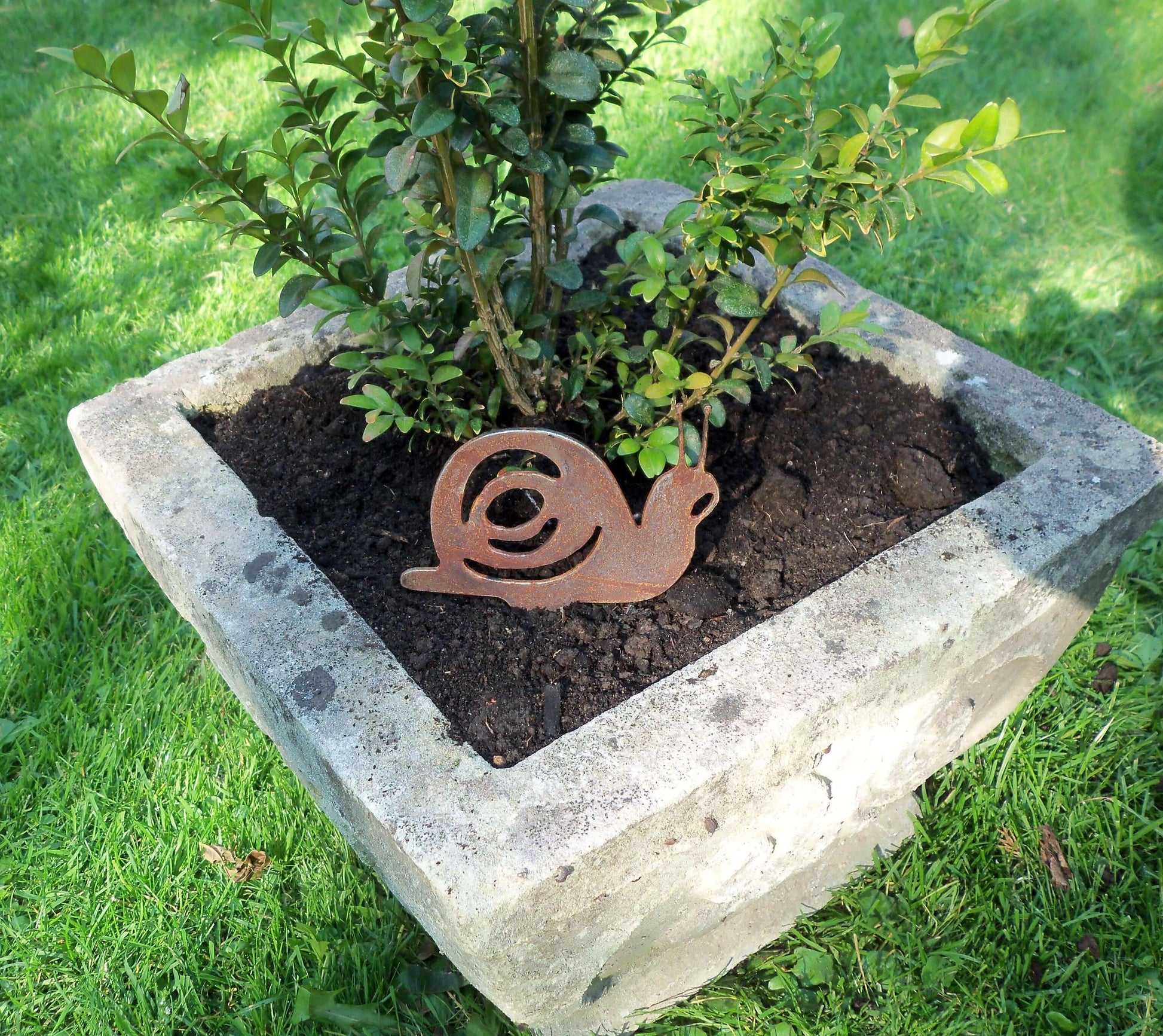 Rusty metal snail garden decoration with pot spike in a stone pot beside green plant, unique patina effect.