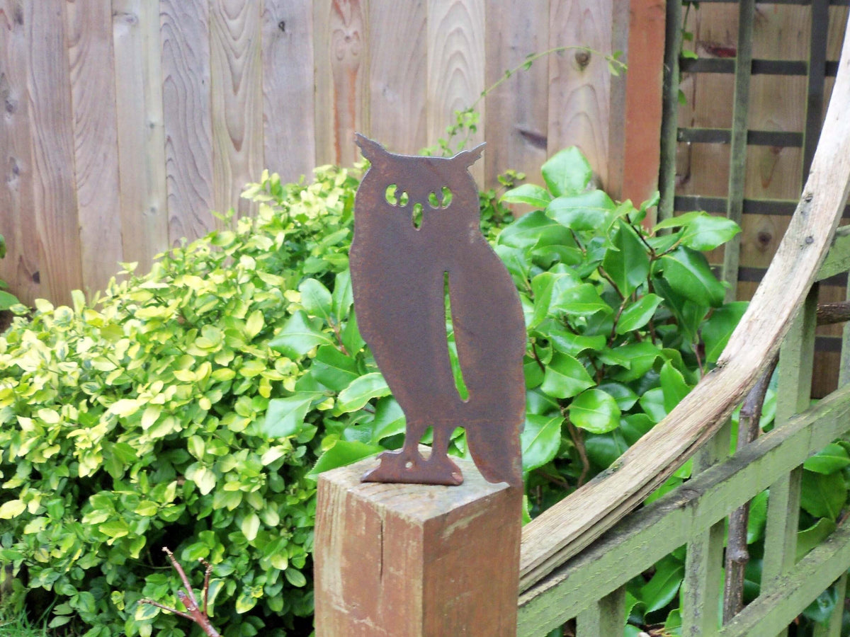 Rusted Steel Owl Garden Ornament atop a wooden post surrounded by lush greenery, showcasing rustic charm.
