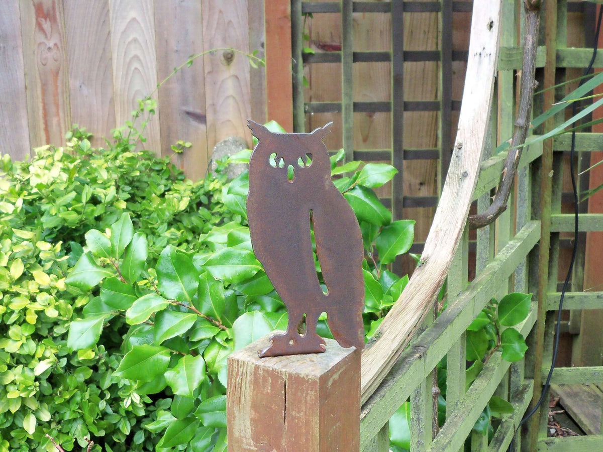 Rusted Steel Owl Garden Ornament perched on a wooden fence post, surrounded by green foliage, adding rustic charm.