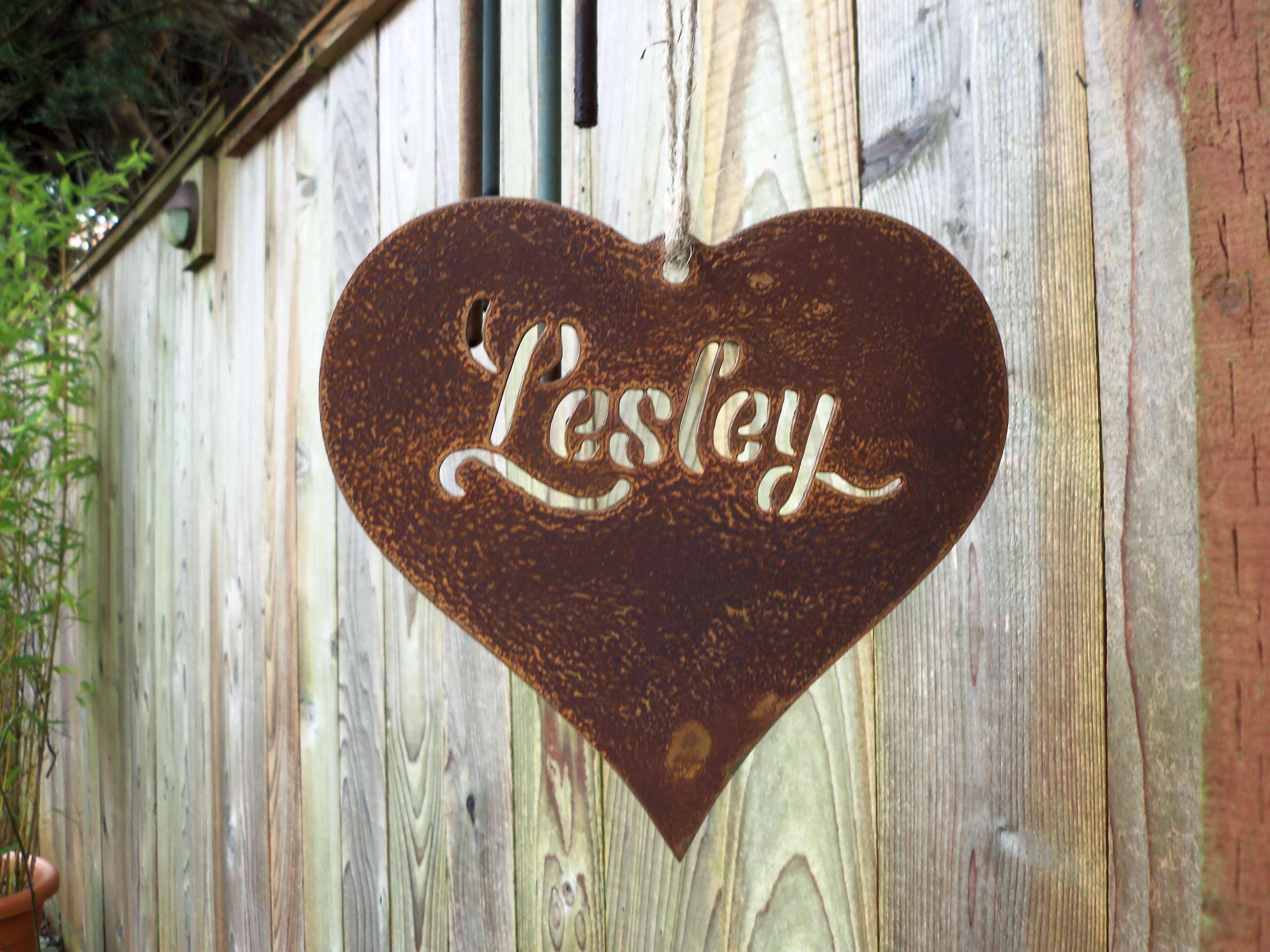 Personalised metal hanging heart garden ornament with a rusty finish & the name 'Lesley' displayed on a wooden background.