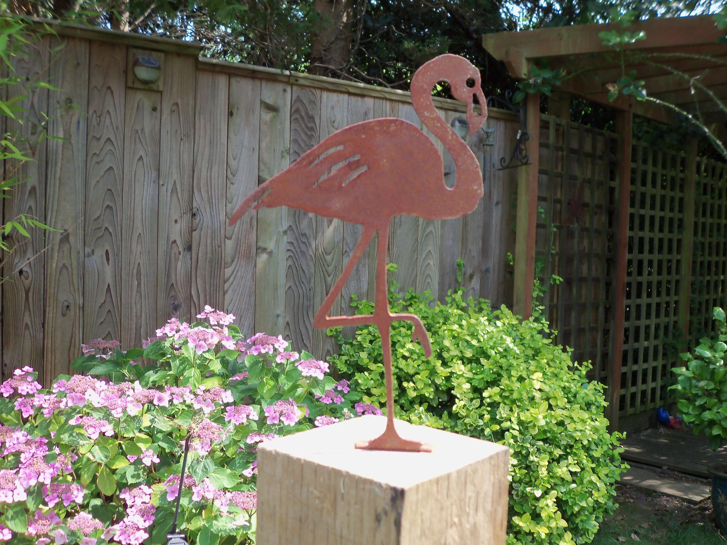 Rusty Metal Flamingo garden ornament on a wooden post surrounded by colourful flowers in a garden setting.