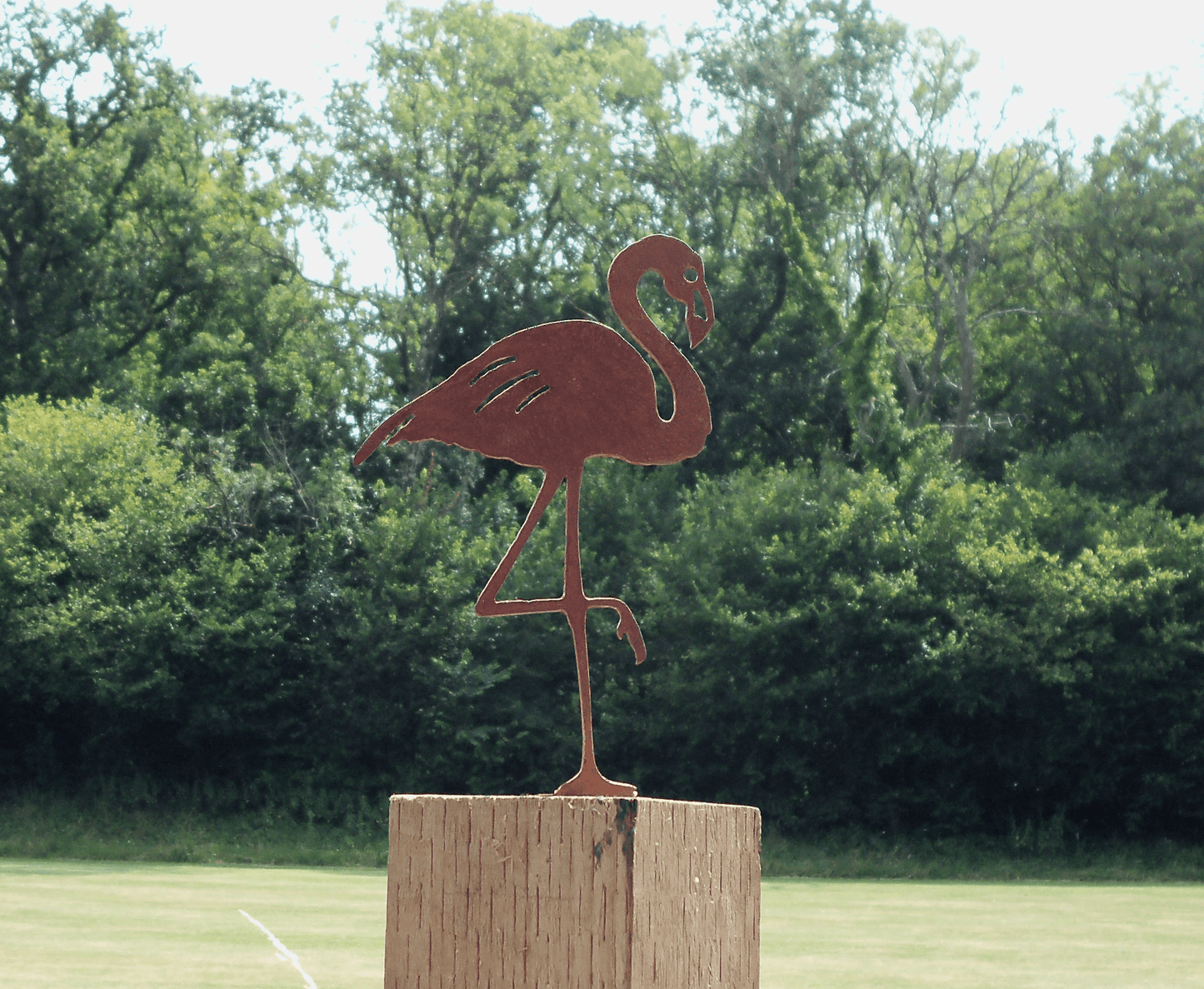 Rusty Metal Flamingo garden ornament standing on a wooden post in a garden surrounded by greenery.