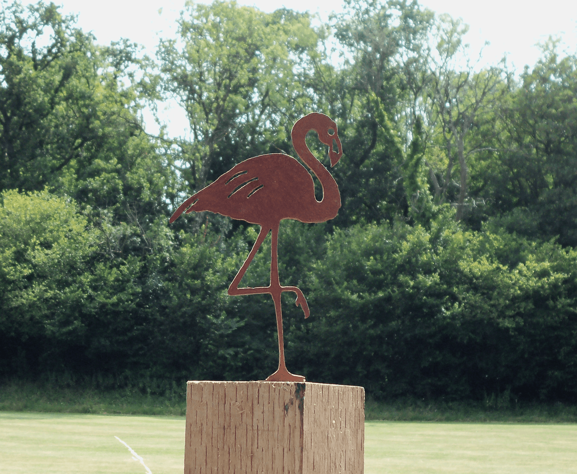 Rusty Metal Flamingo garden ornament standing on a wooden post in a garden surrounded by greenery.