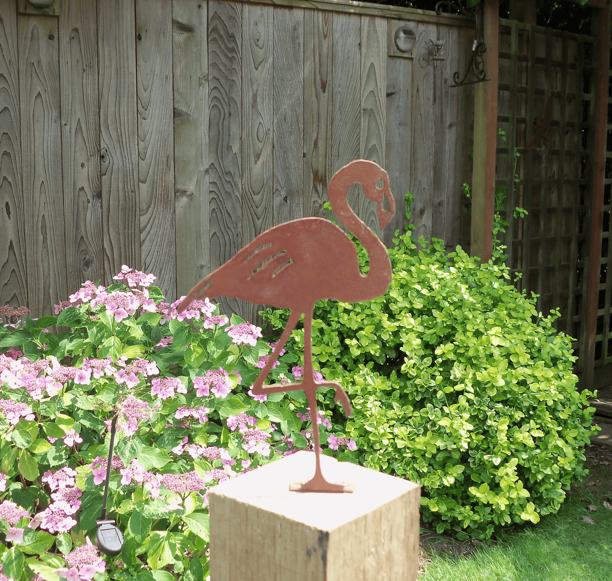 Rusty Metal Flamingo garden ornament displayed in garden with flowers and greenery.