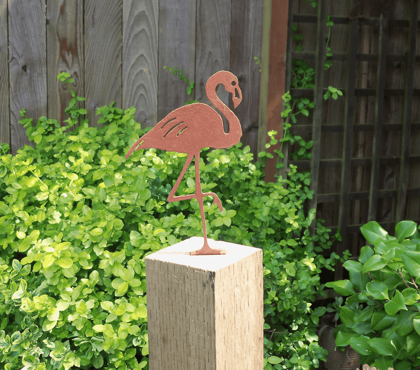 Rusty Metal Flamingo garden ornament atop a wooden post surrounded by green foliage.