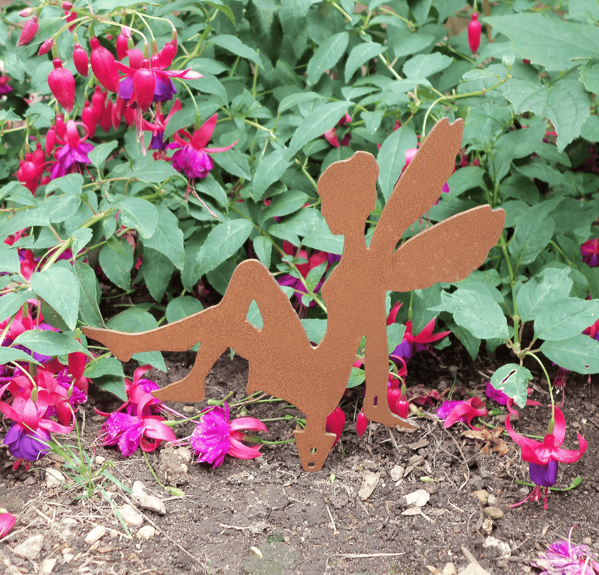 A rusty steel fairy garden ornament with ground spike displayed outdoors in a garden bed among green foliage and pink fuchsia flowers.