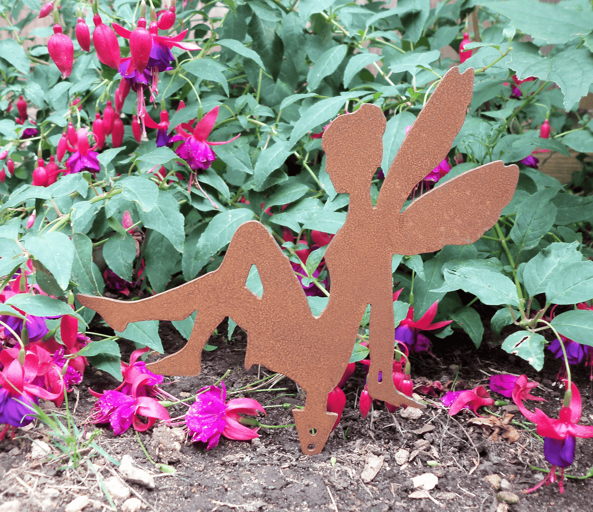 A rusty steel fairy garden ornament with ground spike, displayed outdoors in a garden bed among green foliage and pink fuchsia flowers.