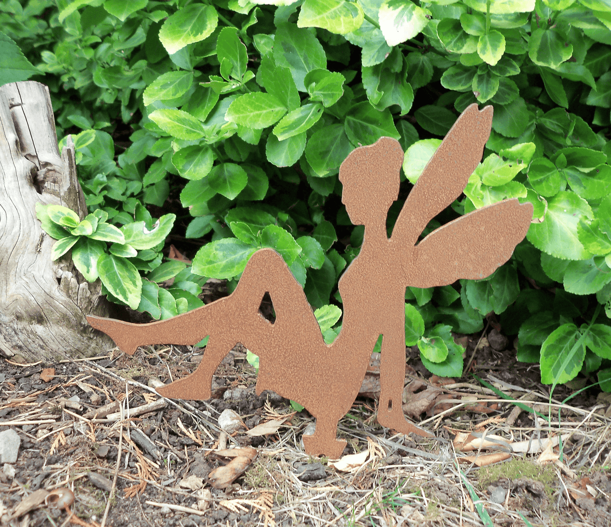 A rusty steel fairy garden ornament with ground spike sitting on the ground, outdoors in front of dense green foliage.