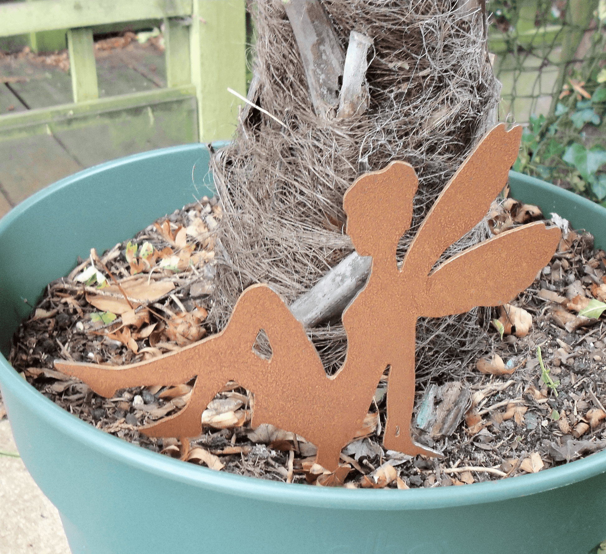 Rusty steel fairy garden ornament with pot spike, and a small tree, displayed in a green planter filled with soil and mulch outdoors.