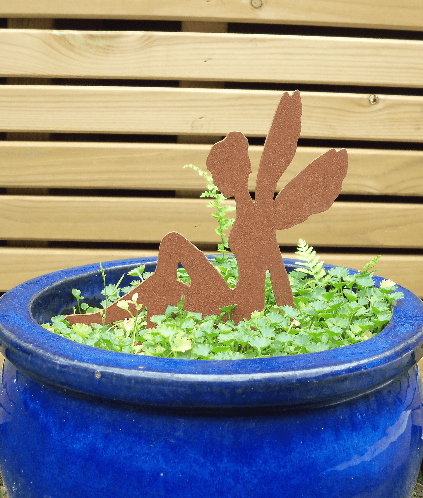 Rusty steel fairy garden ornament with pot spike sitting among green plants in a glossy blue ceramic pot, set against a light wood slat background.