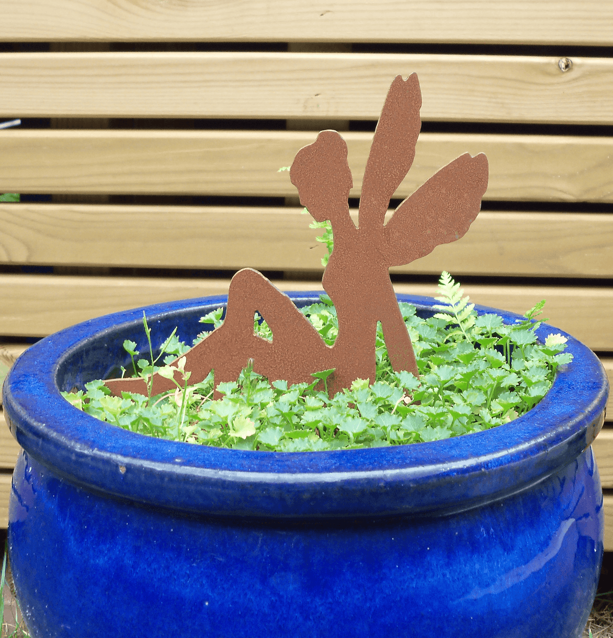 A rusty steel fairy garden ornament with pot spike in green foliage inside blue ceramic pot, set against a light wood slat background.