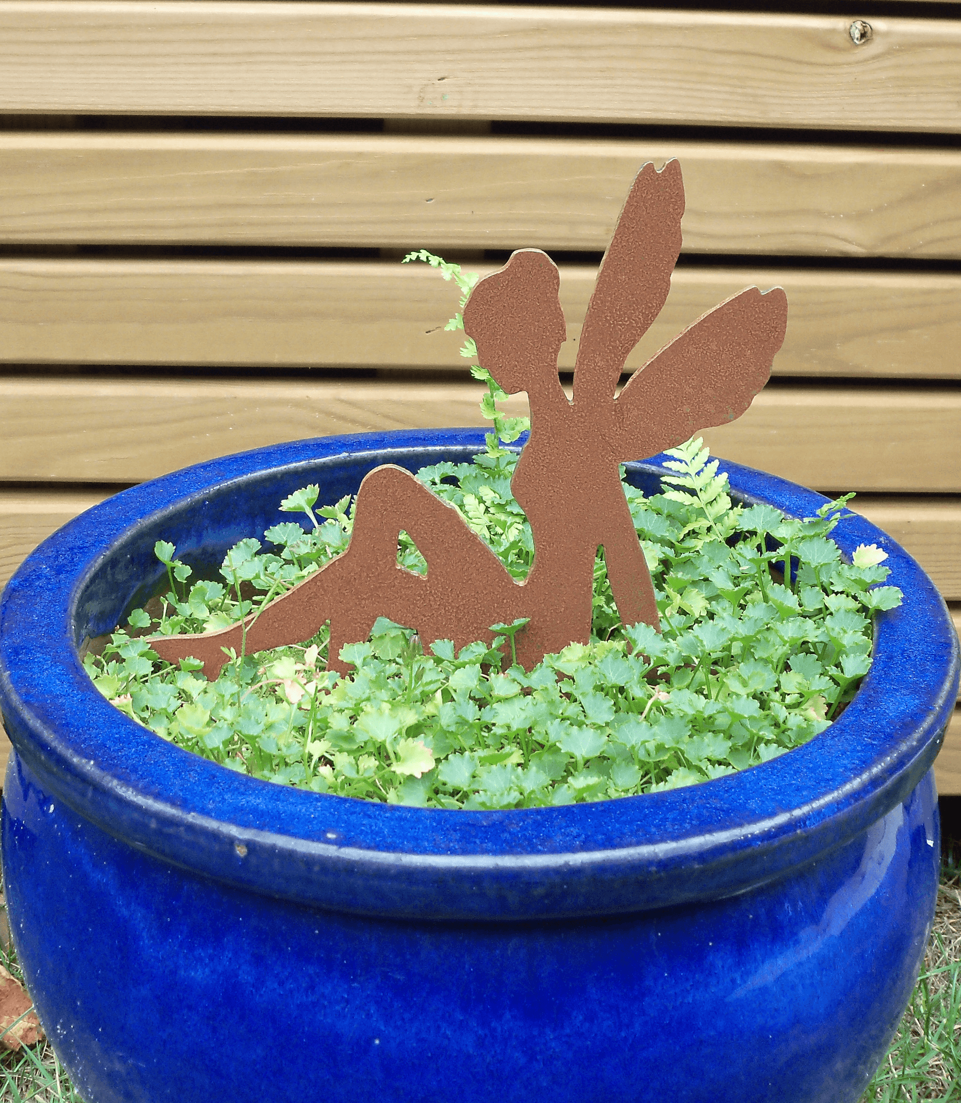 A blue ceramic planter filled with green leafy plants and a rusty steel fairy garden ornament with pot spike, set against a light wood siding background.