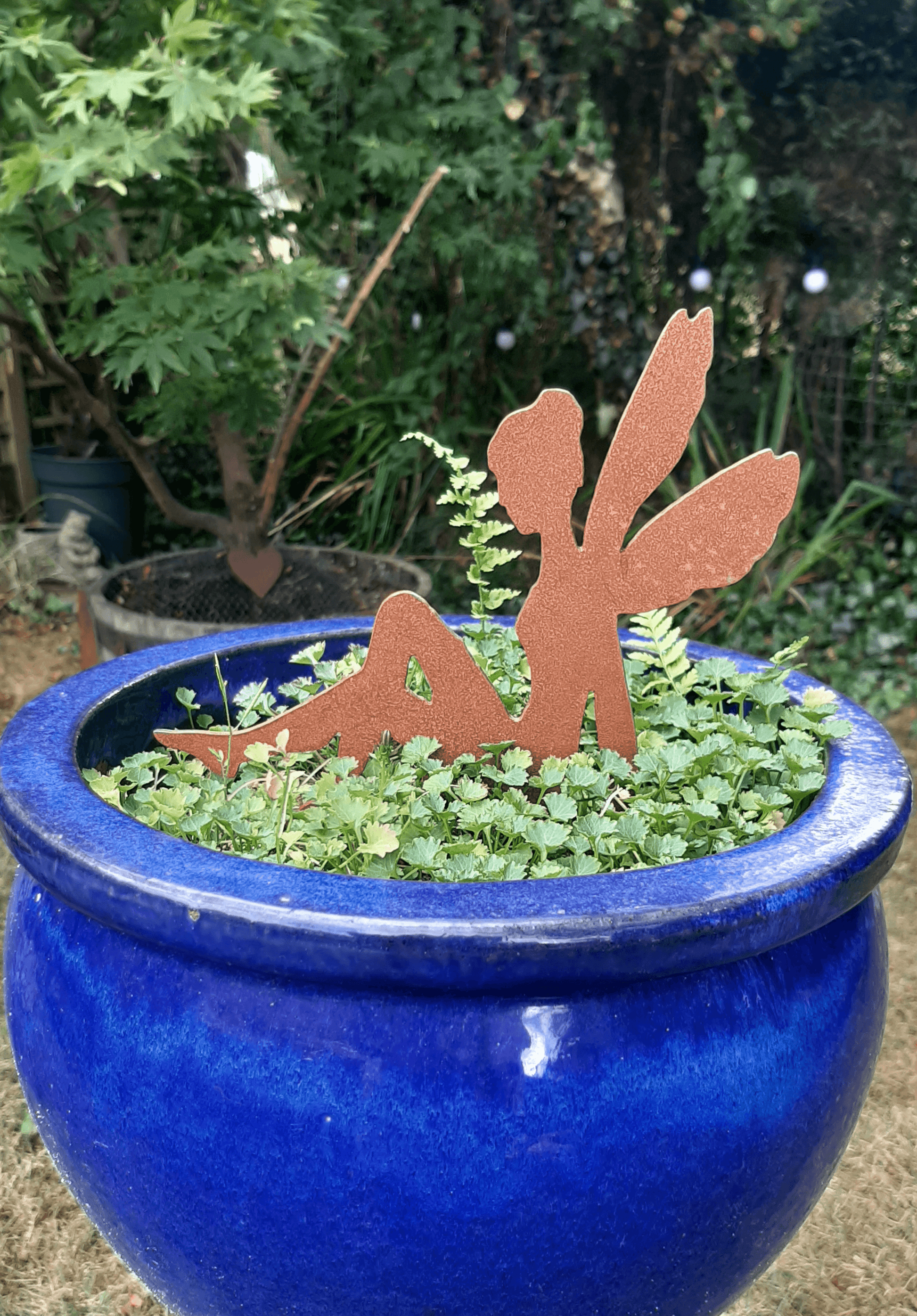 Rusty Steel fairy garden ornament with pot spike in a blue ceramic pot filled with green foliage, set in an outdoor garden background