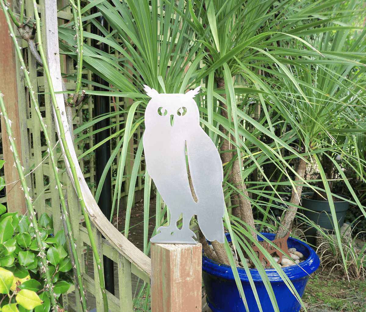 Rustic Steel OWL Garden Ornament displayed on a fence post among lush greenery and potted plants in a garden.