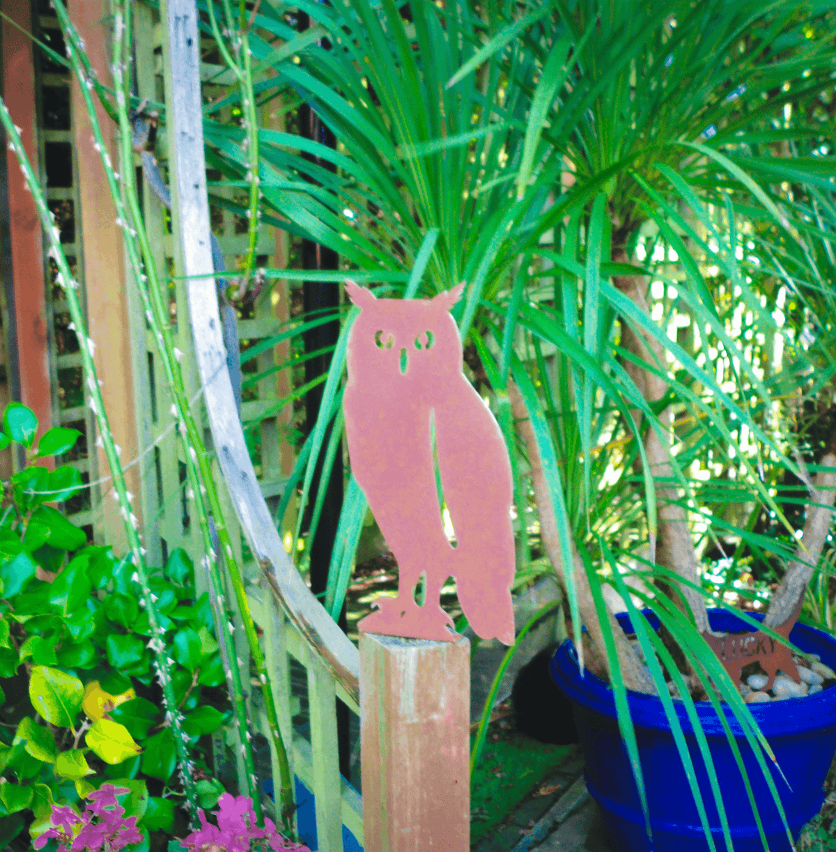 Rustic Steel OWL Garden Ornament placed on a wooden fence post surrounded by lush greenery and potted plants.