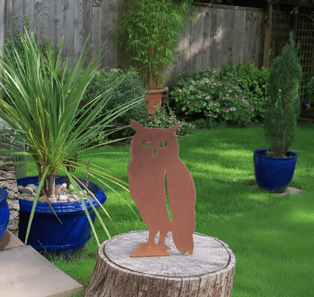 Rustic Steel OWL Garden Ornament displayed in a vibrant garden setting with green grass and colorful potted plants.