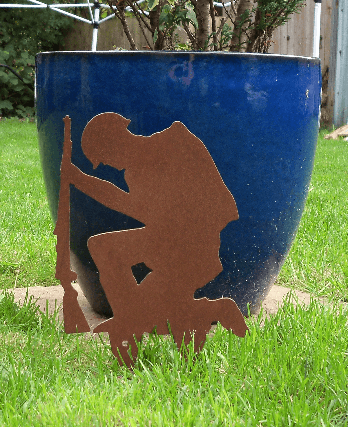 Rustic Steel SOLDIER Garden Ornament positioned near a blue pot in a lush green garden; kneeling soldier with rifle design.