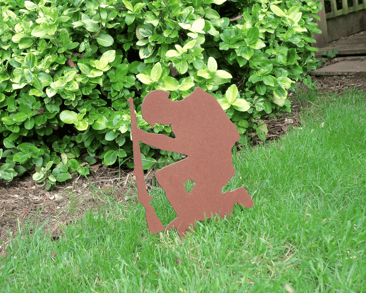 Rustic Steel SOLDIER Garden Ornament displayed in grass with green bushes in the background, featuring a kneeling metal soldier silhouette.