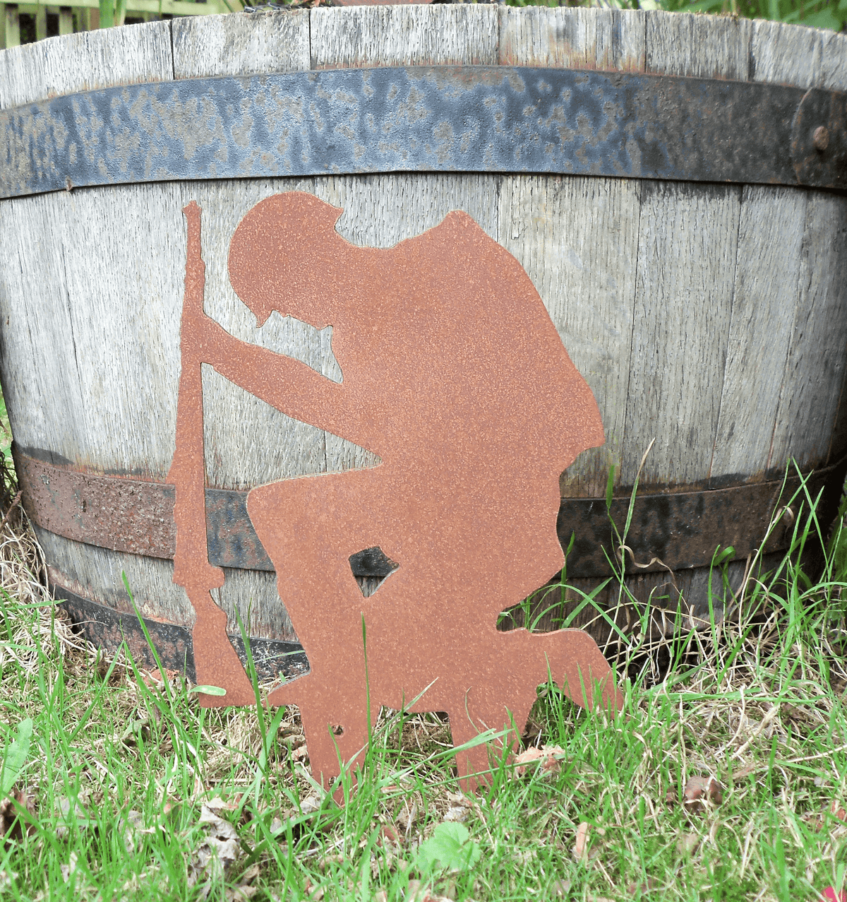 Rustic Steel SOLDIER Garden Ornament displayed next to a rustic wooden barrel in a grassy garden.