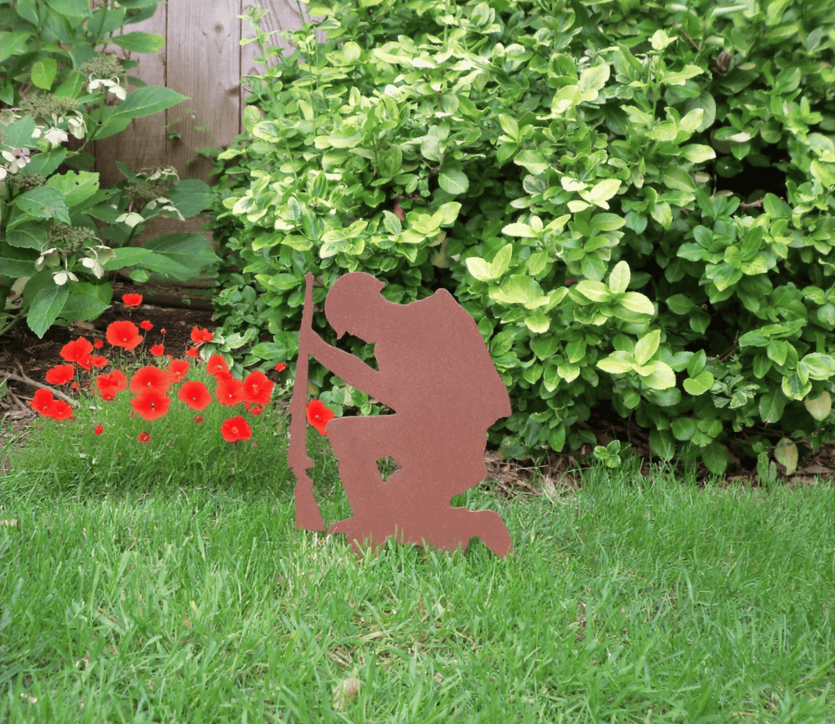 Rustic Steel SOLDIER Garden Ornament set against green grass and vibrant flowers, showcasing a kneeling military figure.