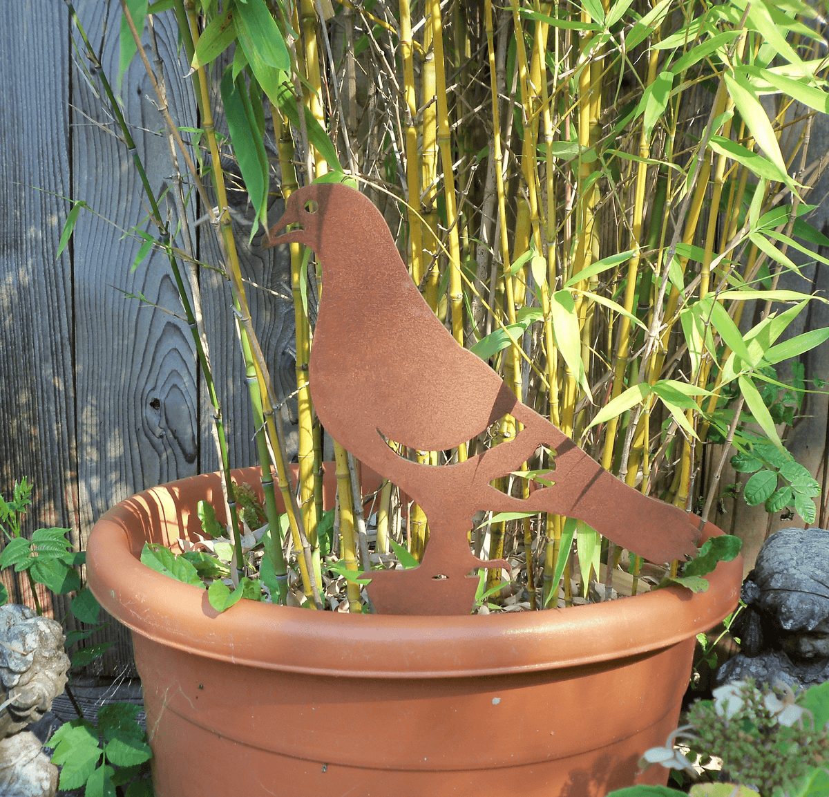 Rusty Steel PIGEON Garden Ornament standing in a flower pot surrounded by bamboo plants, showcasing a rustic design.