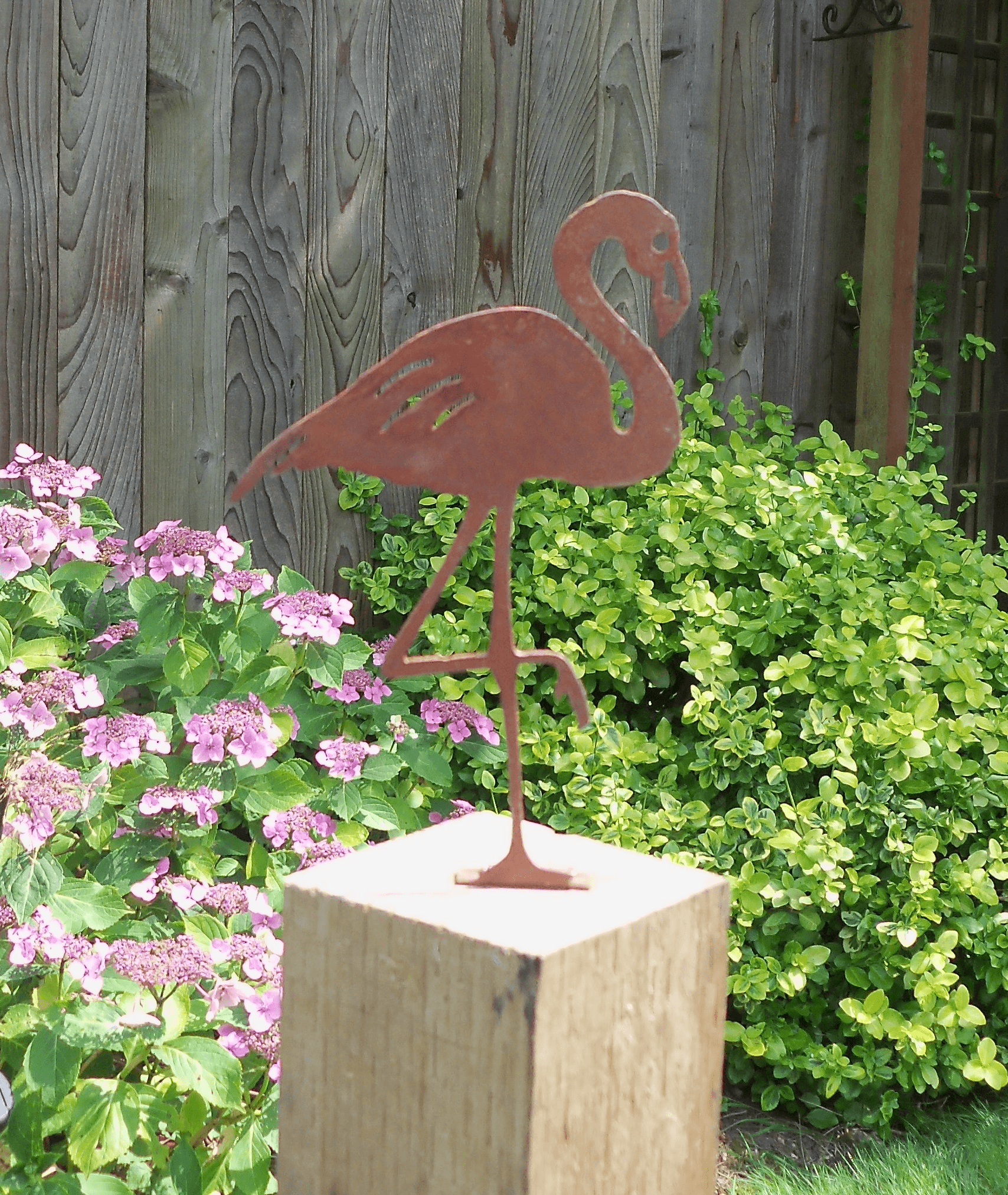 Rusty Metal Flamingo garden ornament atop a wooden post surrounded by blooming flowers in a garden setting.