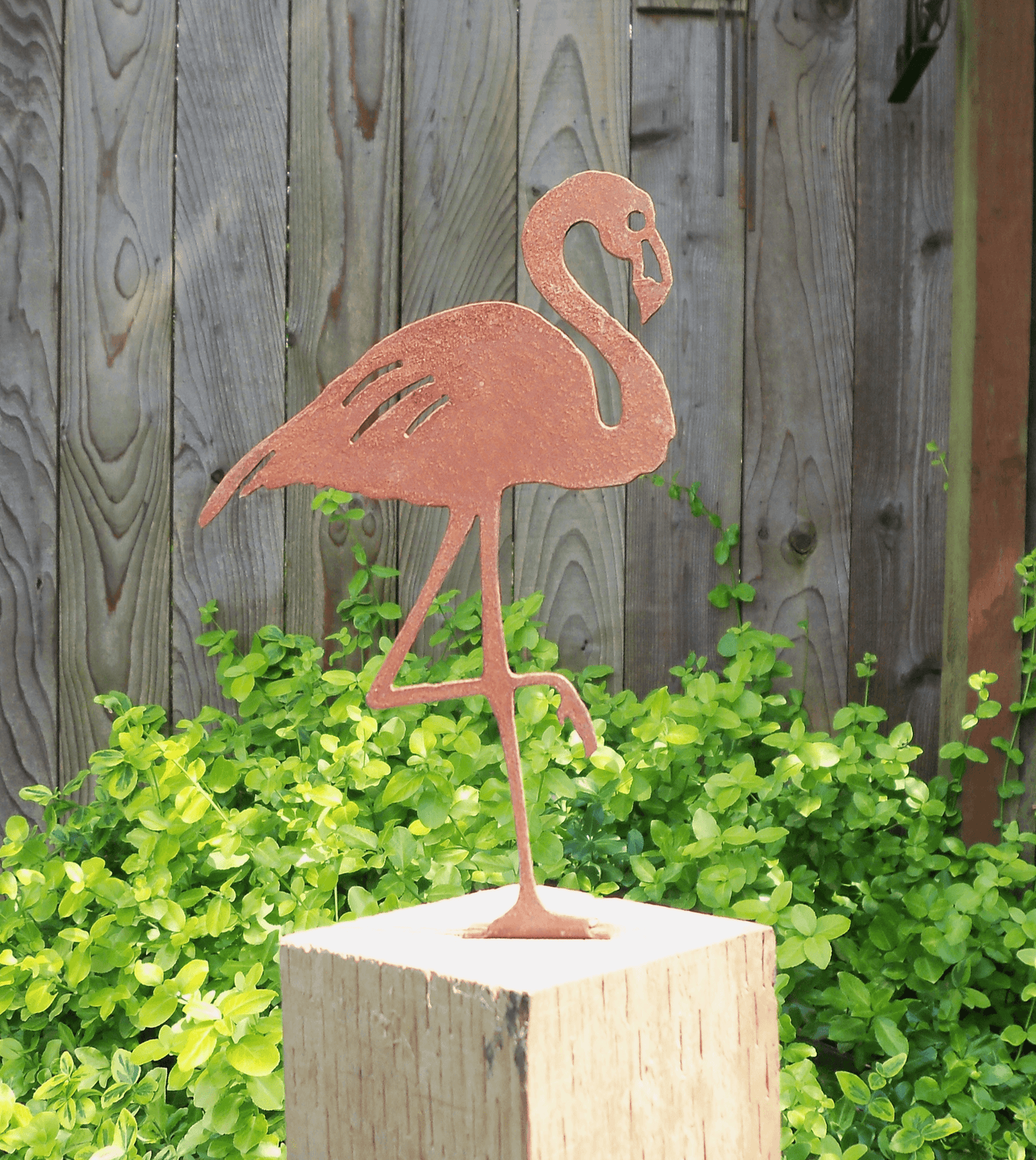 Rusty Metal Flamingo garden ornament on a wooden post surrounded by greenery in a garden setting.