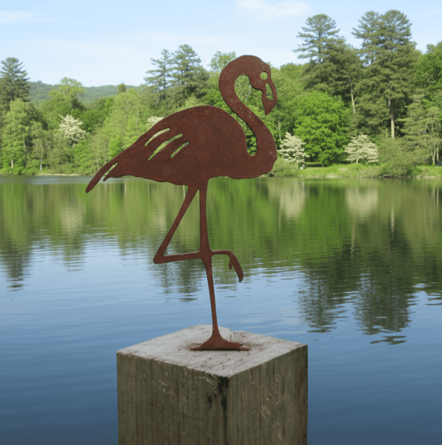 Rusty Metal Flamingo garden ornament on a wooden post by a tranquil lake surrounded by trees.