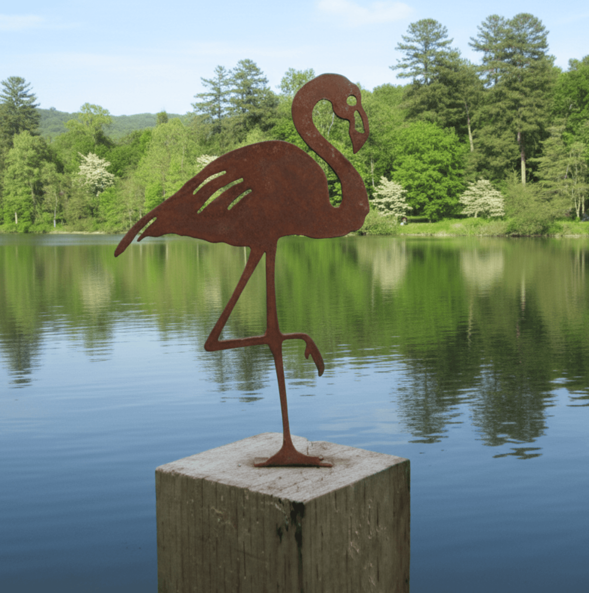 Rusty Metal Flamingo garden ornament on a wooden post by a tranquil lake surrounded by trees.