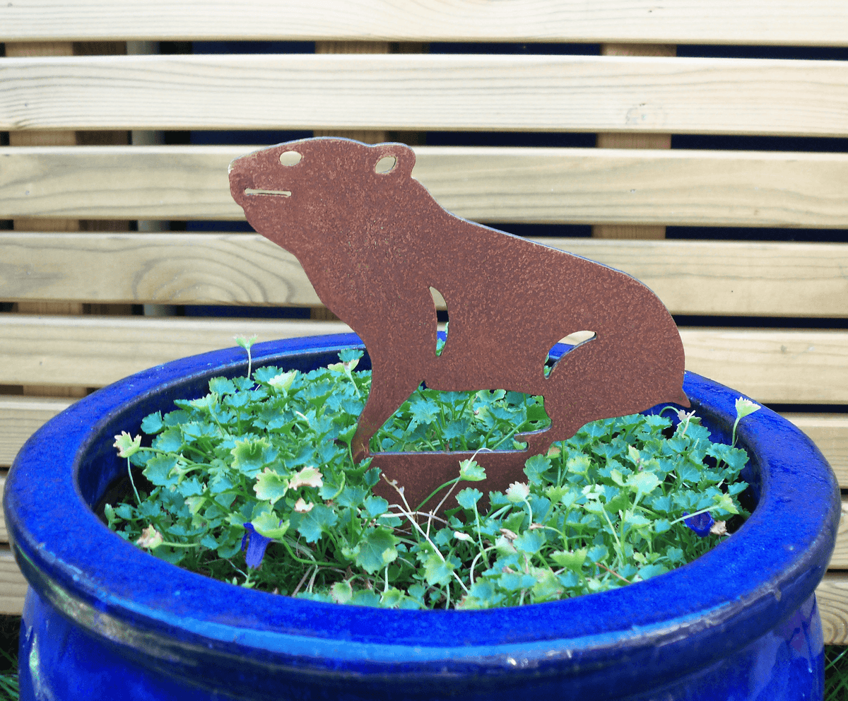 Rusty Steel HAMSTER Garden Ornament in a blue pot surrounded by vibrant green plants, adding rustic charm to outdoor decor.