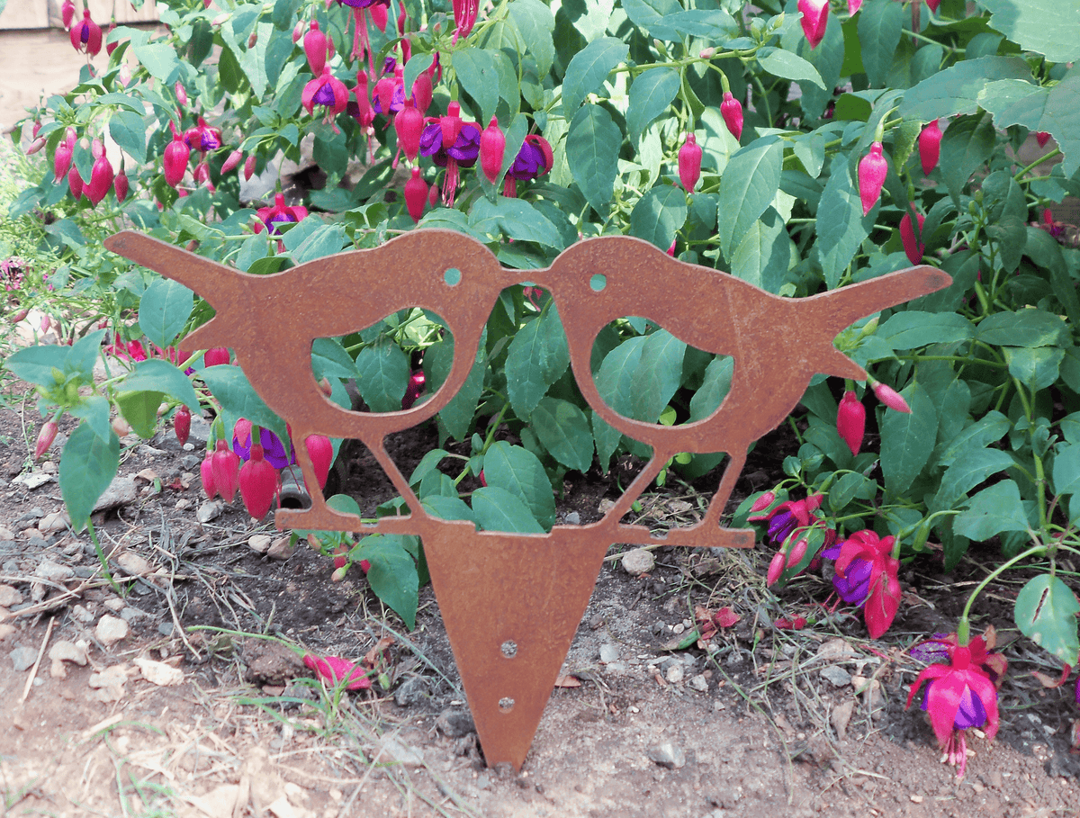 Rusty steel kissing robins garden ornament displayed in a colorful garden with pink flowers.