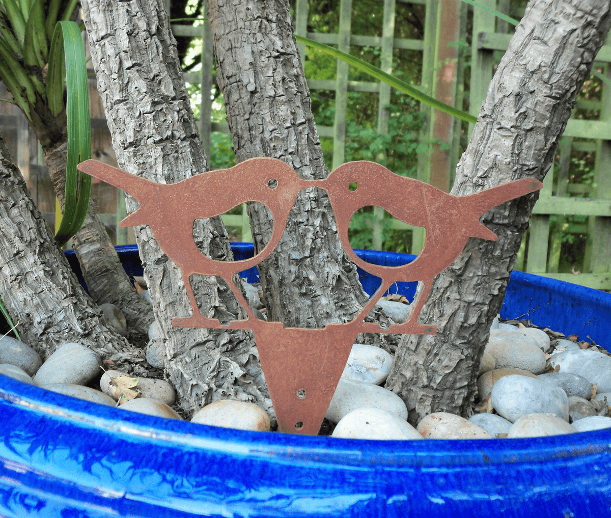 Rusty steel kissing robins garden ornament nestled in a blue pot with pebbles and tree branches.