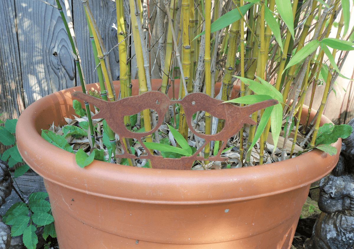 Rusty steel kissing robins garden ornament nestled in a flower pot surrounded by green plants.